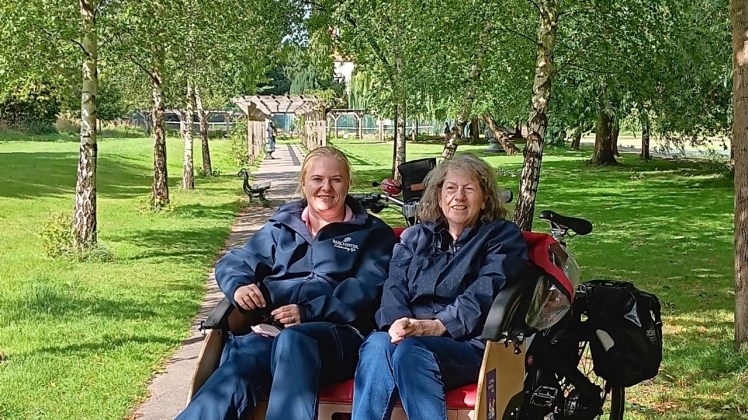 Two people seated in a Triobike under the trees in Rowntree Park