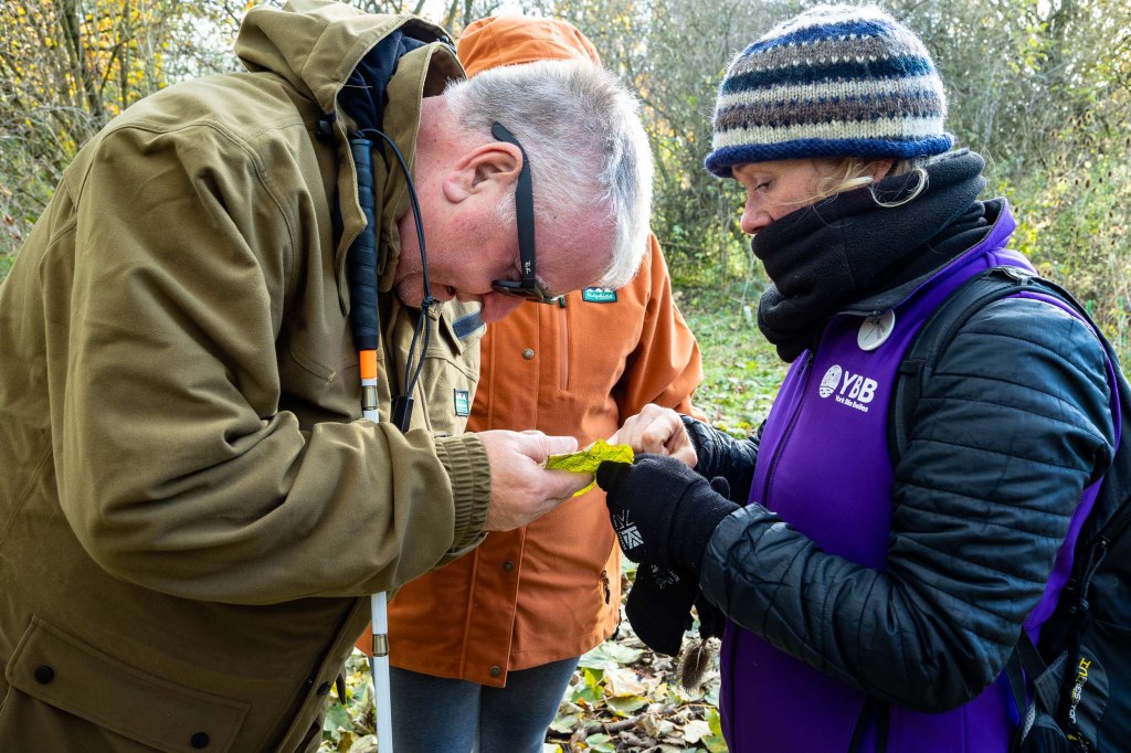 Woman in a YBB gilet is showing the detail on a leaf to a man with a white cane 